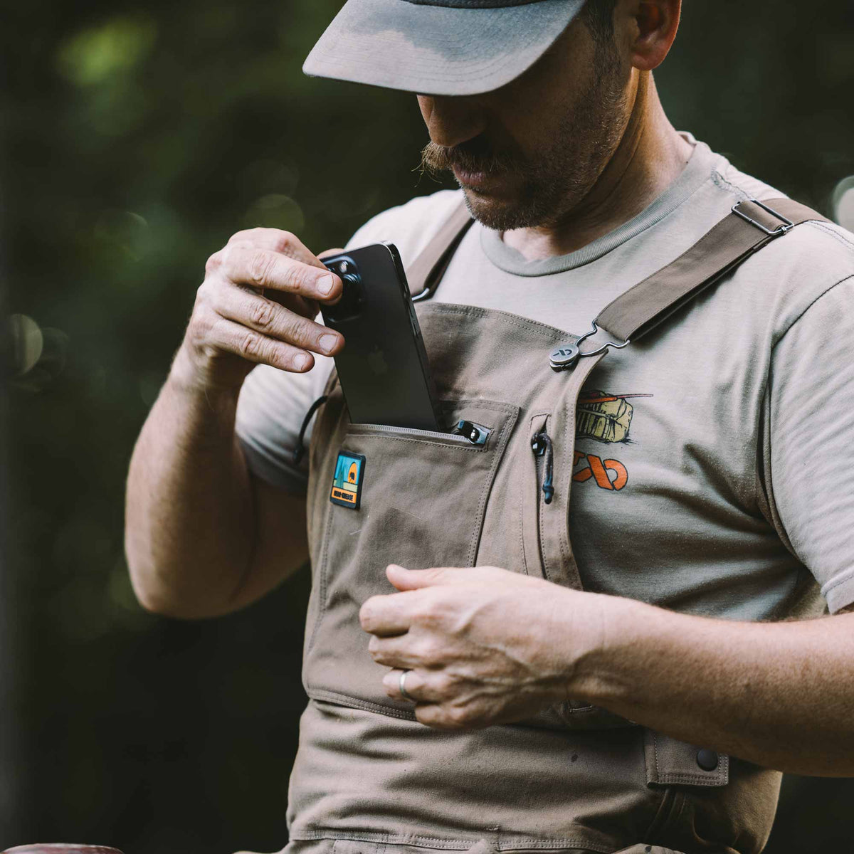 A man wearing tan utility overalls over a gray t-shirt and a gray cap, holding a black smartphone in his right pocket. The overalls feature a chest pocket with a small blue and orange patch, a pen loop, and a small embroidered logo on the left chest.