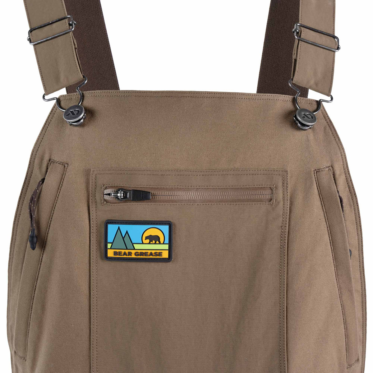 Close-up of tan bib overalls with adjustable shoulder straps, side pockets, and a front zip pocket featuring a 'Bear Grease' patch.
