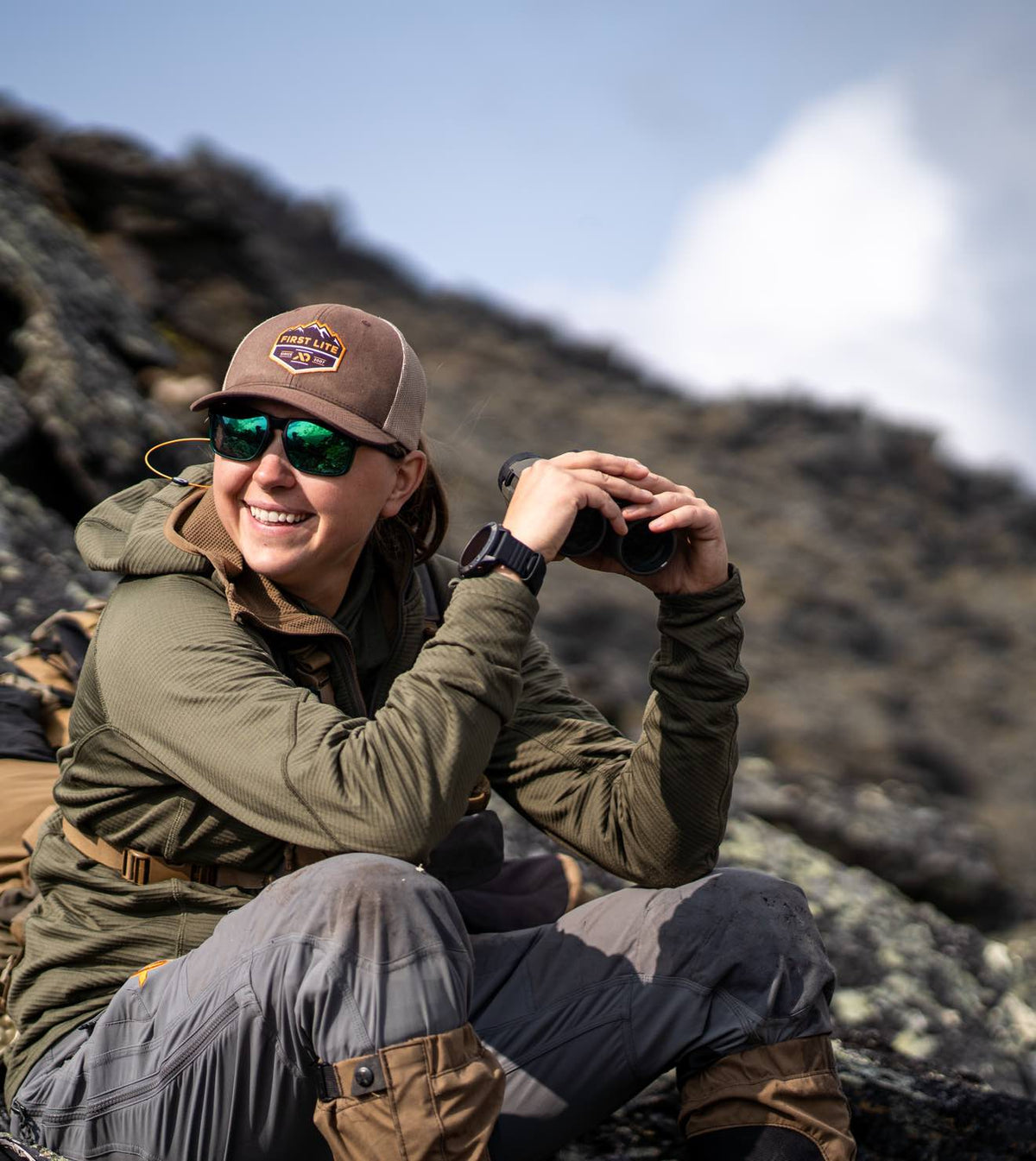 Smiling woman in olive green hoodie, grey pants, brown 'First Lite' hat, green mirrored sunglasses.
