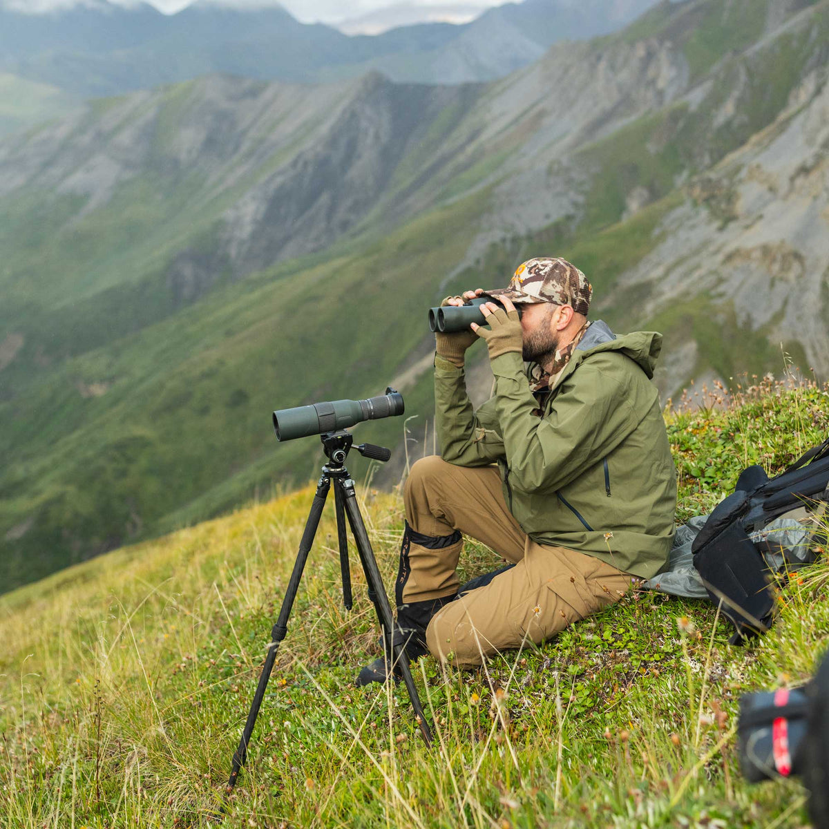 A man in a green rain jacket sits on a grassy mountain slope using binoculars, with a spotting scope on a tripod and a backpack nearby, set against a dramatic green and gray mountain landscape.