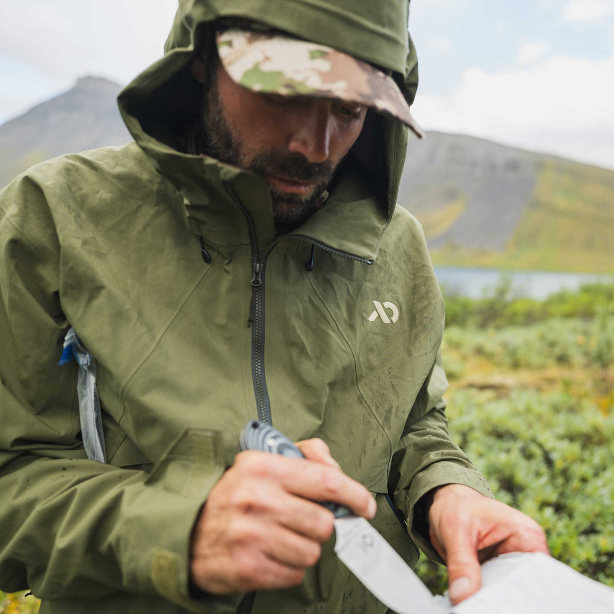 A man wearing an olive green waterproof hooded jacket and a camouflage cap, outdoors near a lake and mountains, holding a knife and paper.