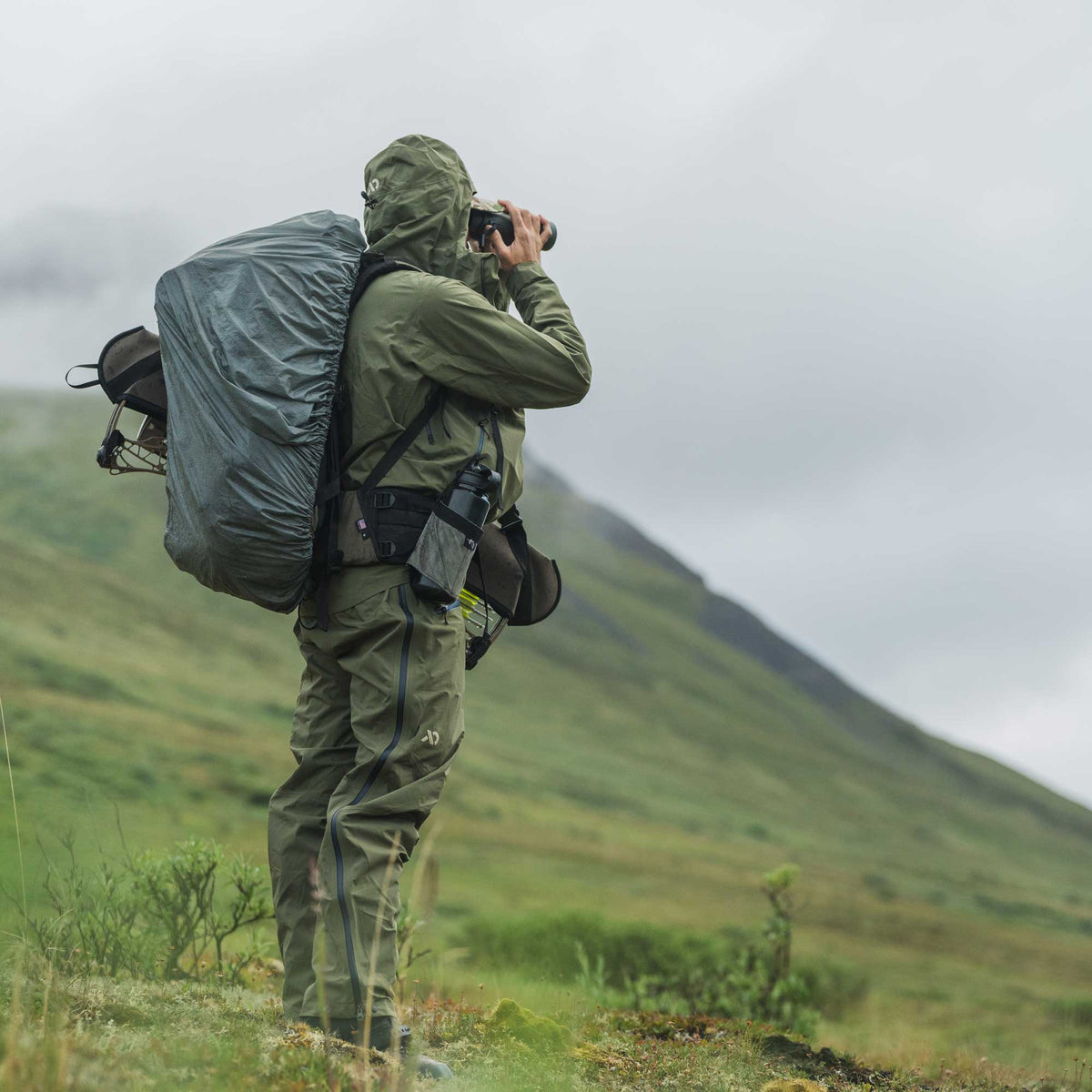 A person in an olive green waterproof suit stands in a grassy mountain landscape under a cloudy sky, holding binoculars and carrying a large gray backpack.