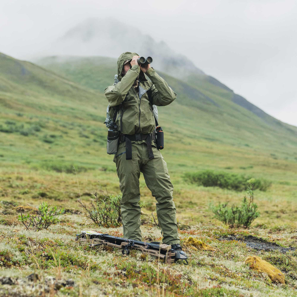 A person in an olive green outdoor hunting suit stands on a grassy mountain slope using binoculars, with a rifle resting on the ground in front of them and misty hills in the background.