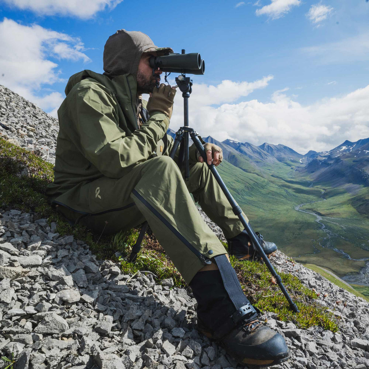 A man in an olive green rain jacket and pants sits on a rocky mountainside using a spotting scope mounted on a tripod, with a vast green valley and mountains under a blue sky in the background.