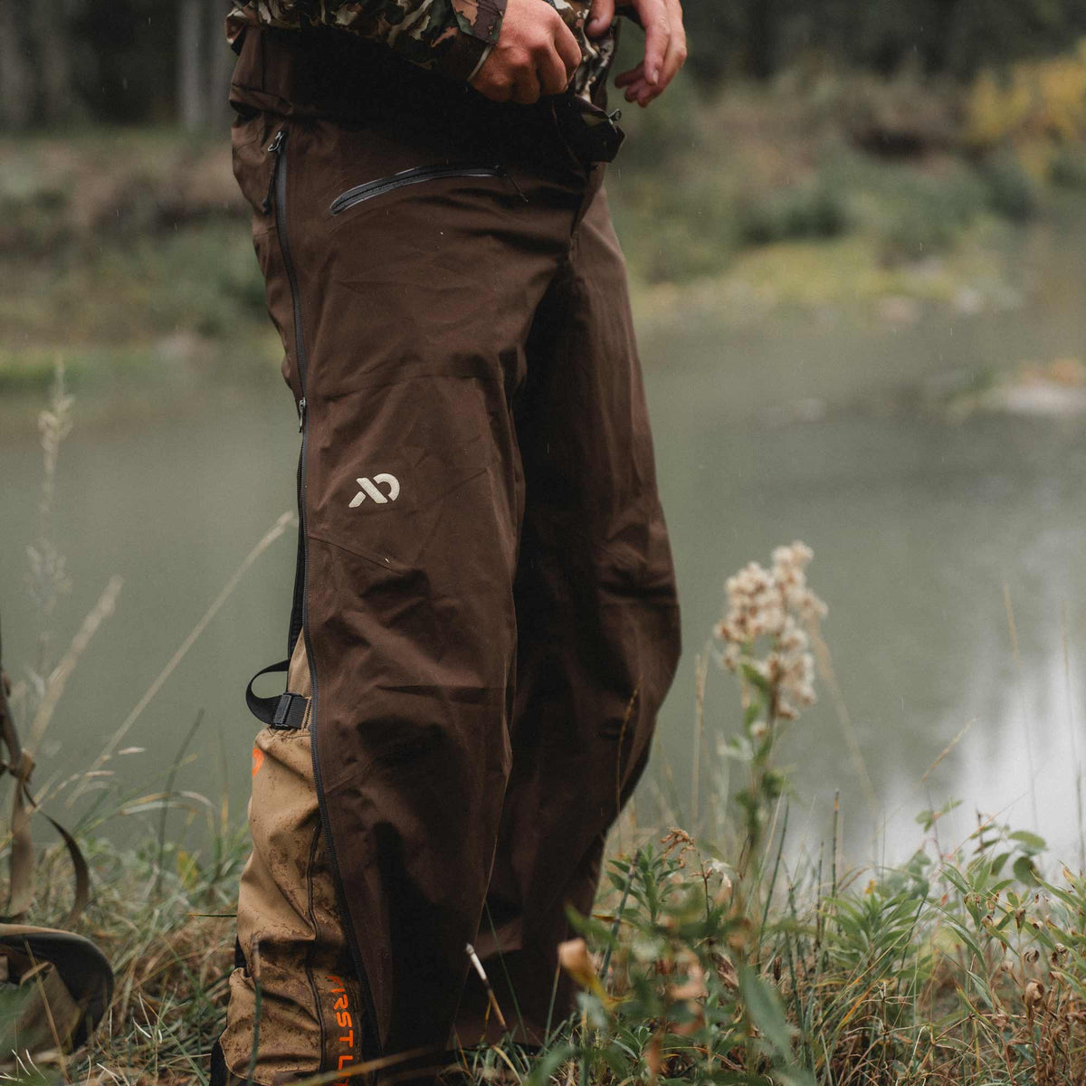 A person outdoors by a body of water wearing brown waterproof rain pants with a visible white logo on the thigh and a tan boot in the same pants, photographed in a natural outdoor setting.