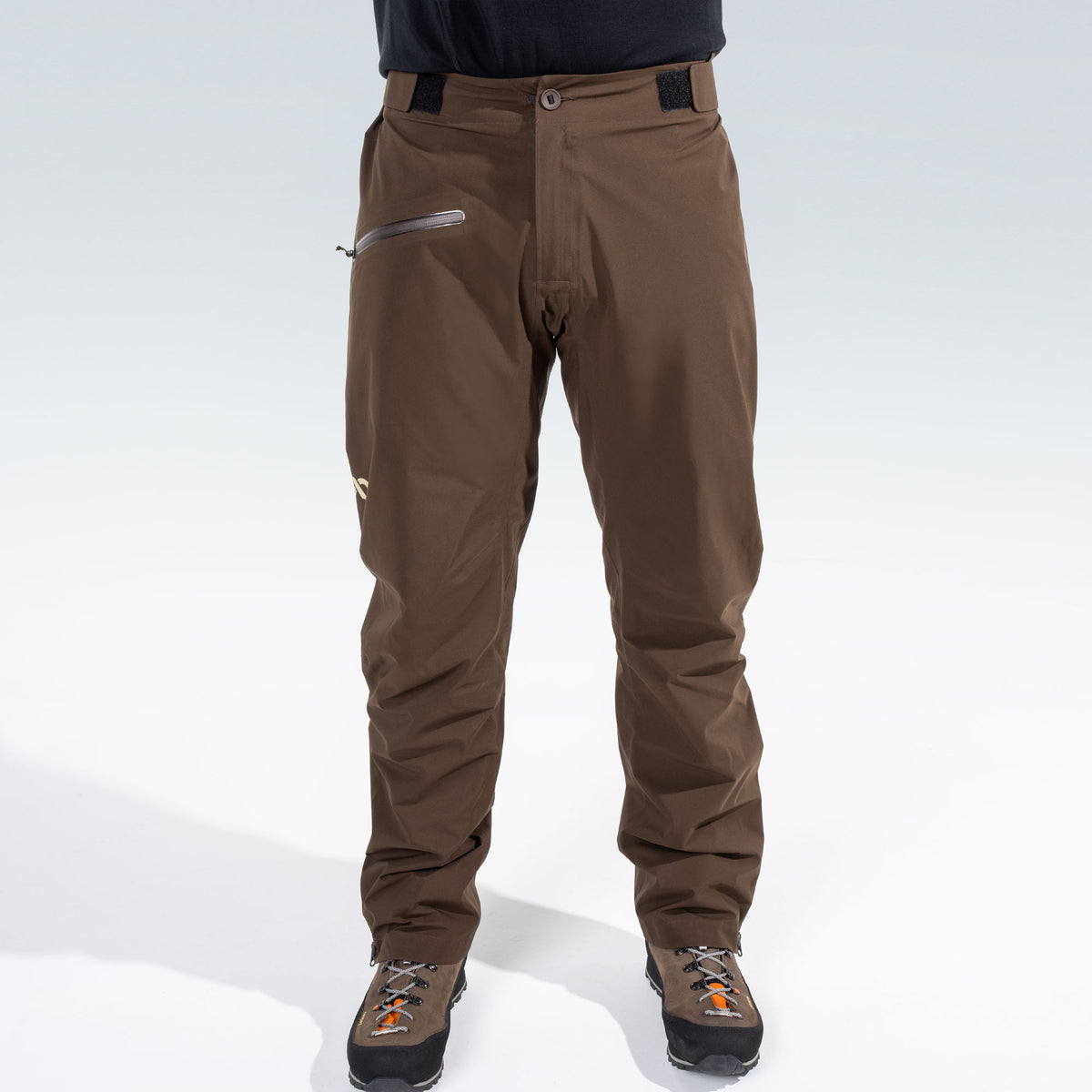 A person from the waist down wearing brown technical rain pants with a zippered pocket and brown hunting boots, shown against a light gray studio background.