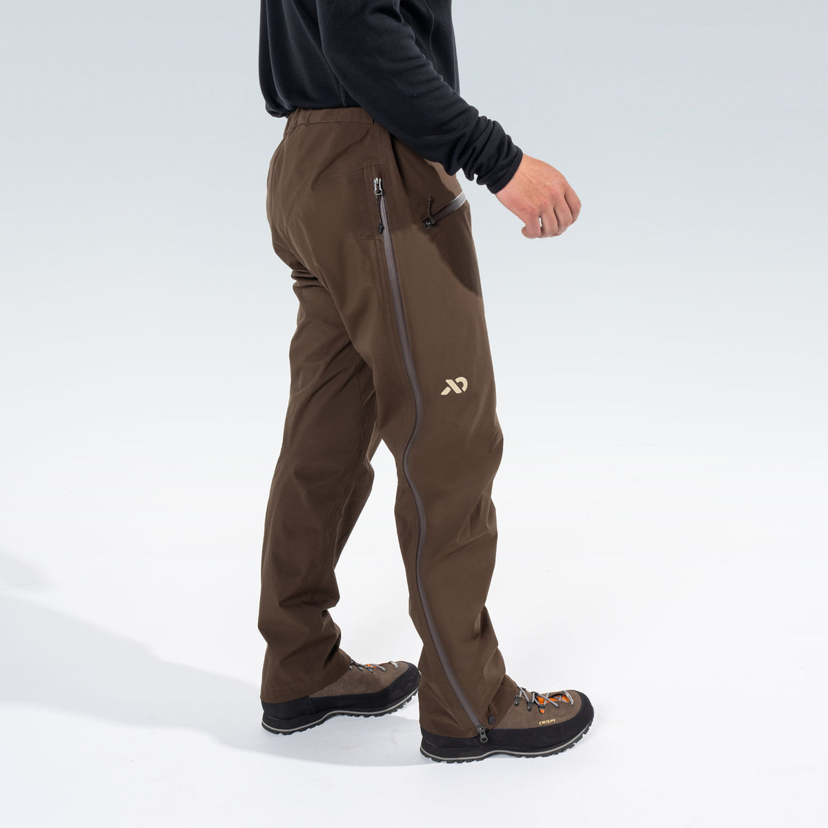 A person wearing brown technical pants with gray zippers and a small tan First Lite logo, shown from the waist down against a clean white studio background.