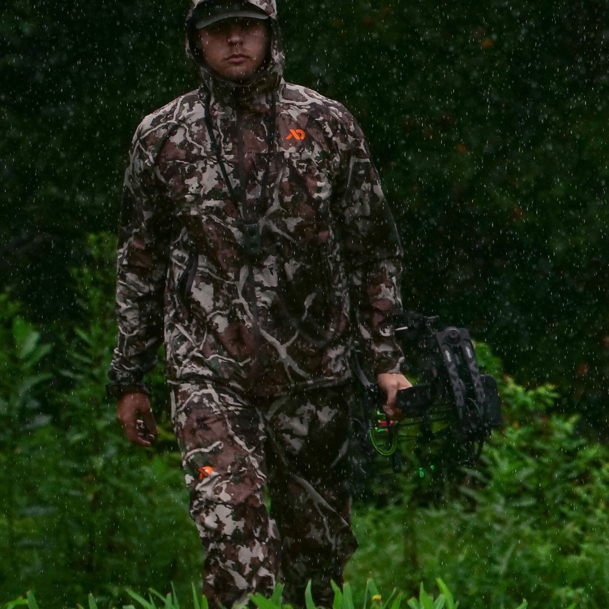 Hunter wearing a brown and grey camouflage rain suit with orange logos, holding a compound bow in the rain.