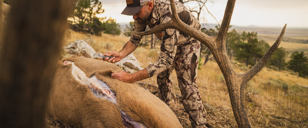 Hunter in camouflage clothing processing an elk in a field with trees and open sky in the background.