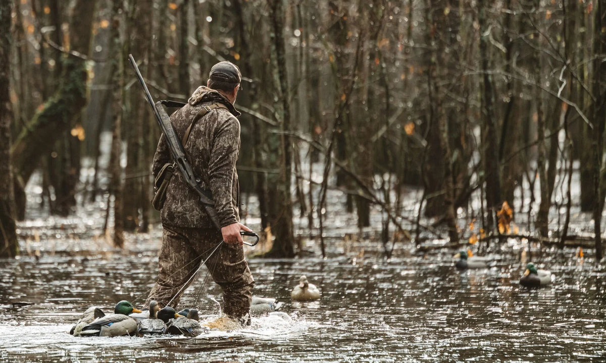 Camo hunter with shotgun and duck decoys wading in flooded woods.