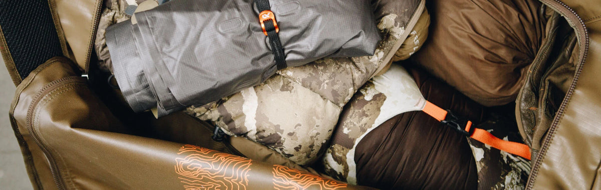 Open olive green duffel bag packed with gray gear, camo items, and orange topo map design.