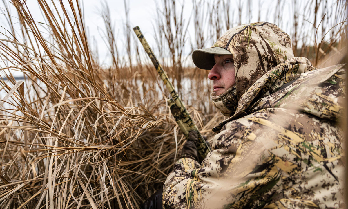 Hunter in camo jacket, cap, and balaclava, holding a camo shotgun in reeds.