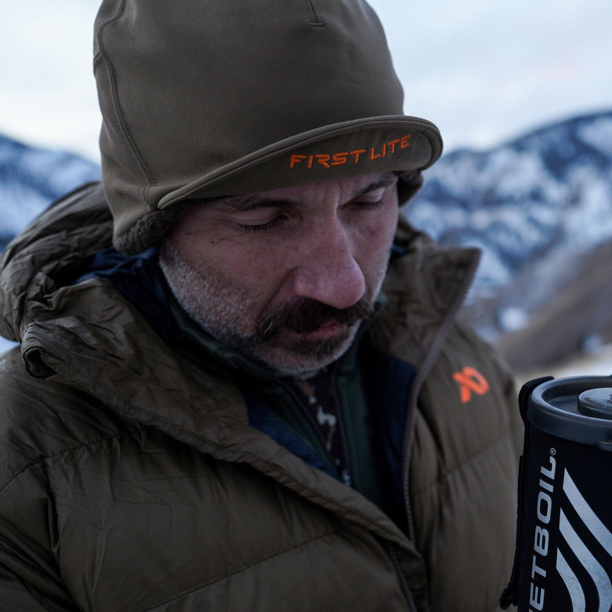 Olive green First Lite winter hat with orange embroidered logo on the brim and visible ear flaps.