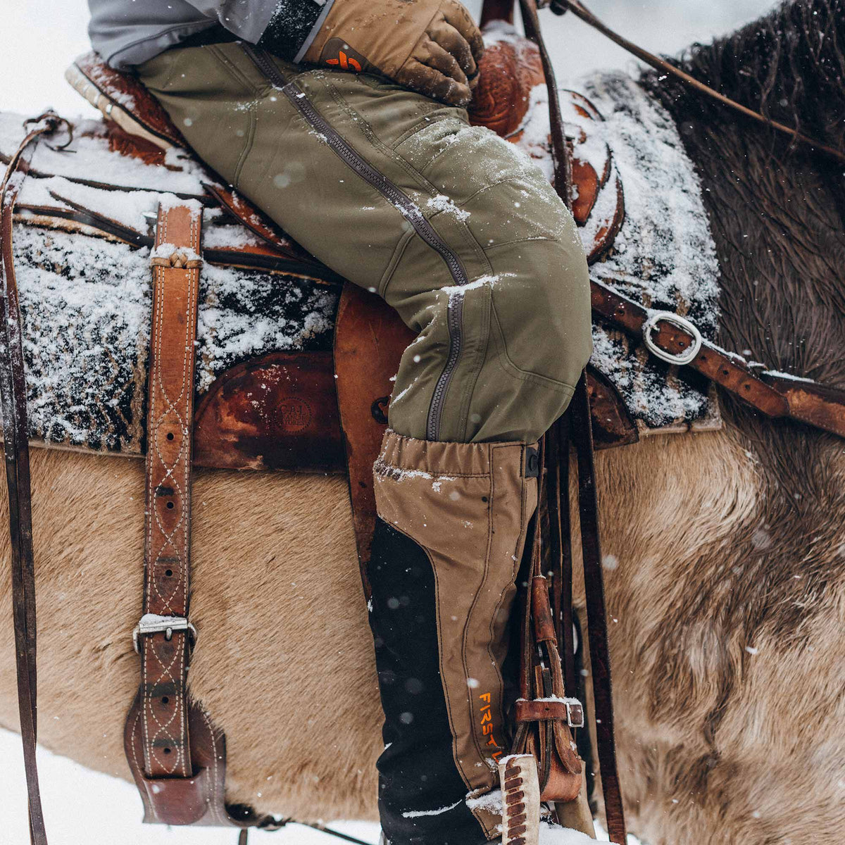 Olive green pants with full-length side zippers, tan lower leg panels, and black reinforced shins featuring the "FIRST LITE" logo.