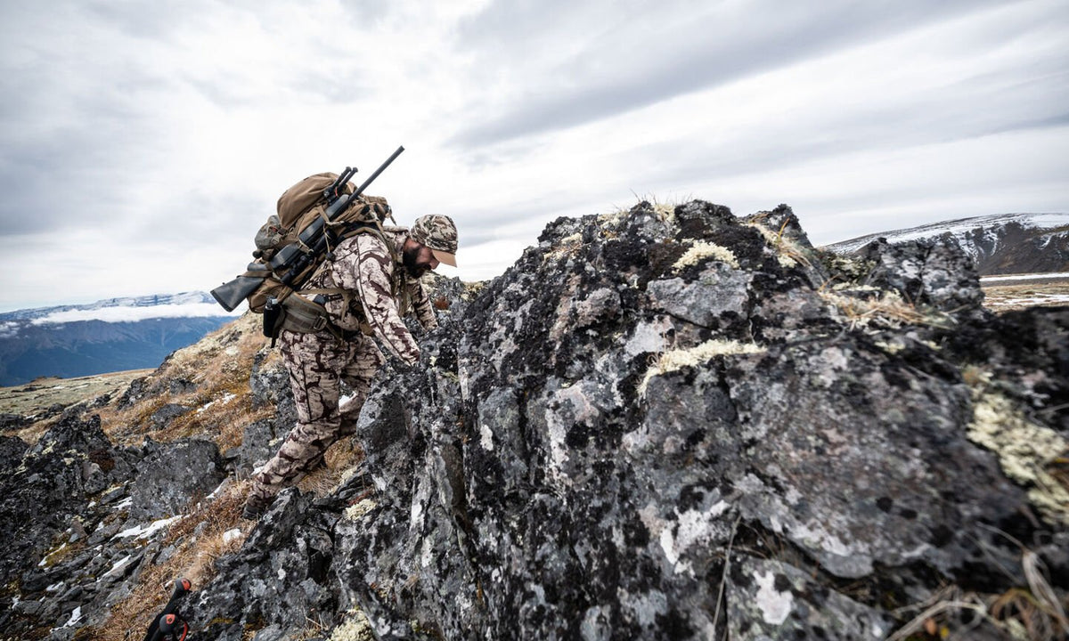 Man wearing tan camouflage hunting gear with rifle and backpack, climbing rocks.