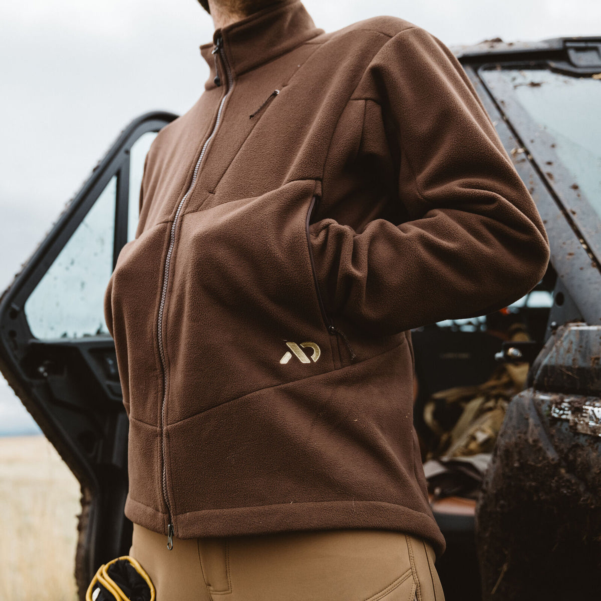 A brown full-zip fleece jacket with a stand-up collar, vertical chest zipper pocket, two side zipper pockets, and a gold logo on the left chest.