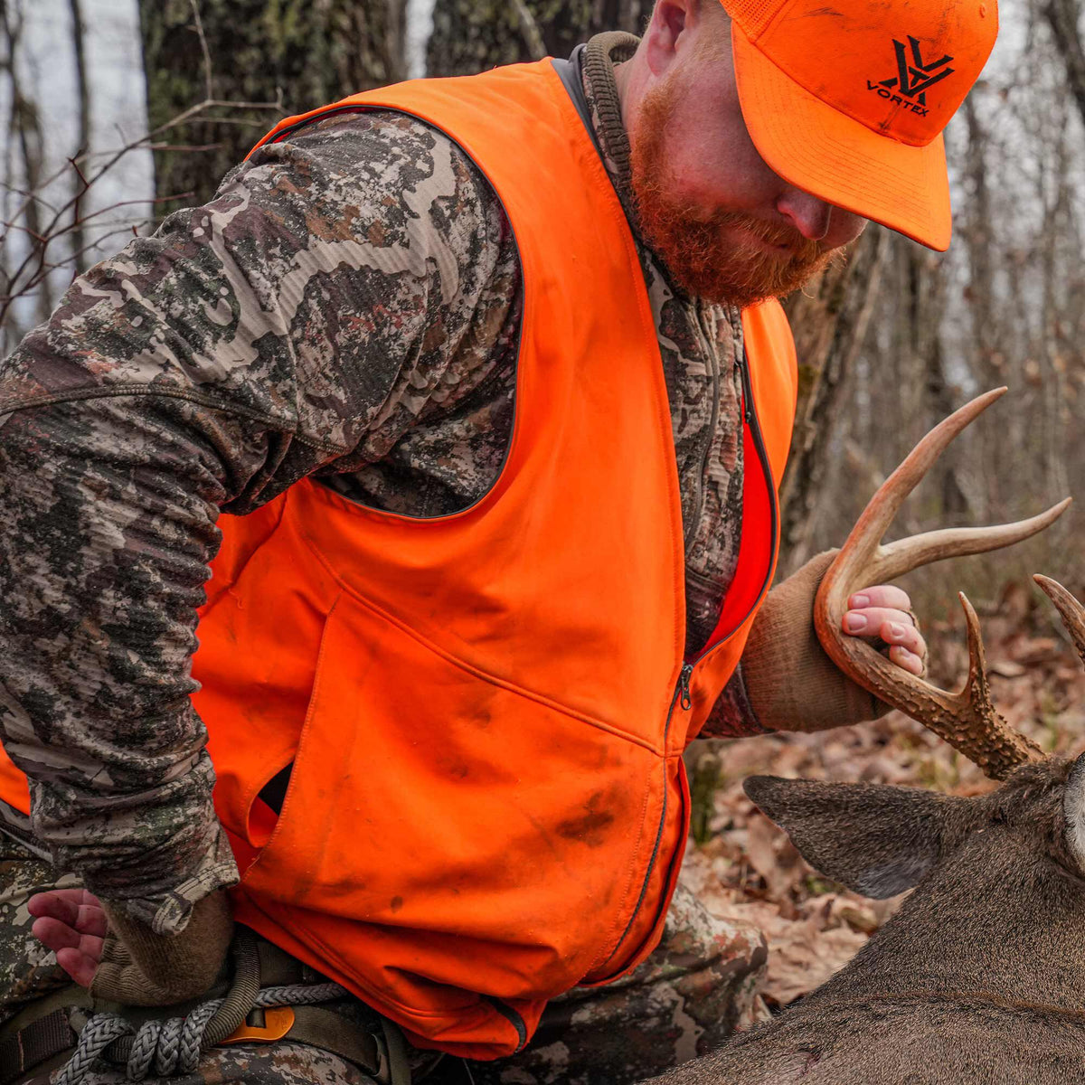 Hunter wearing a blaze orange hunting vest and Vortex logo cap, kneeling beside a deer.