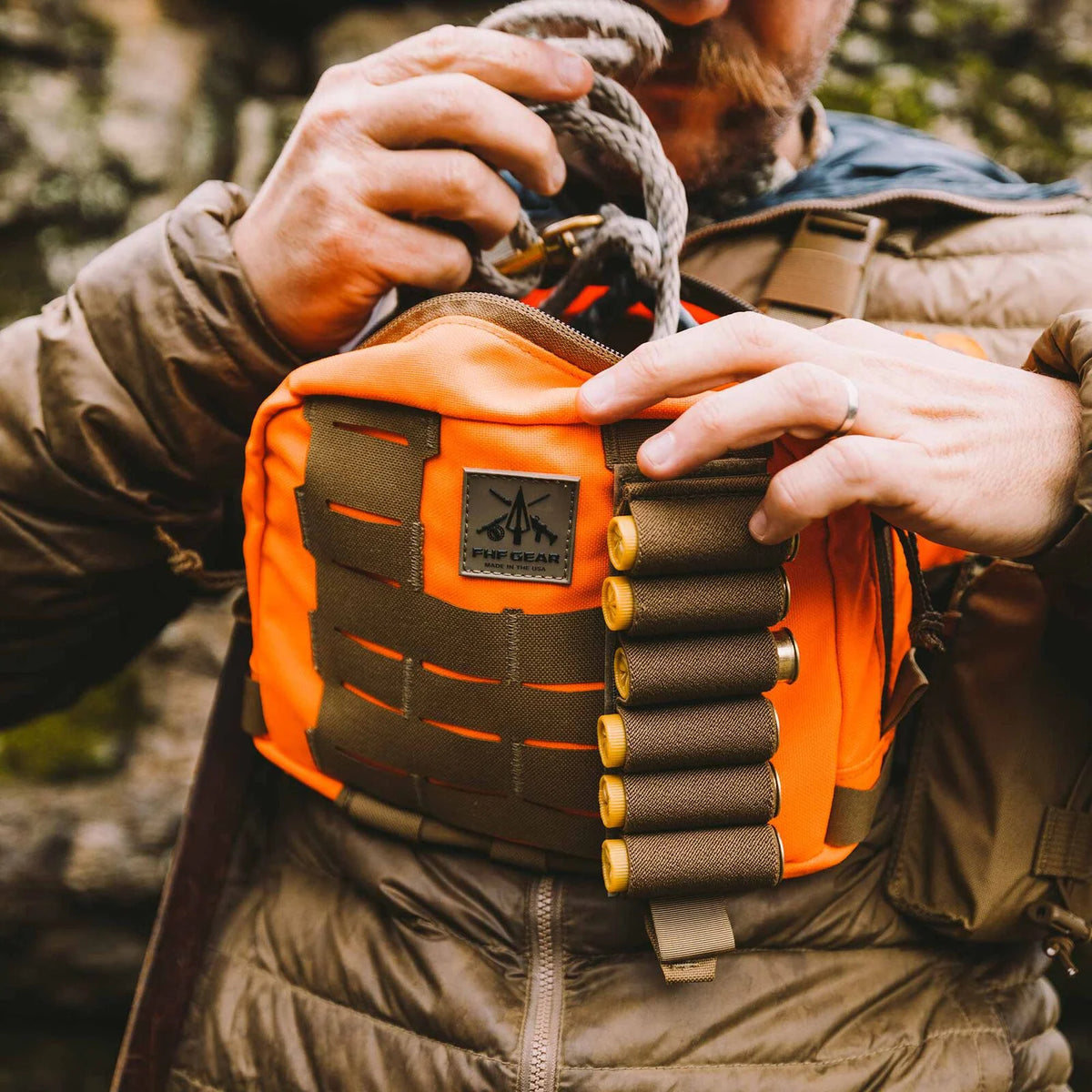 A person accesses an orange and olive FHF GEAR chest pack featuring MOLLE-style webbing and a five-round shotgun shell holder.