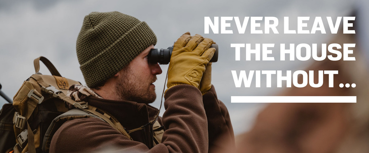 Man looks through black binoculars, wearing olive beanie, tan gloves, and tan backpack.