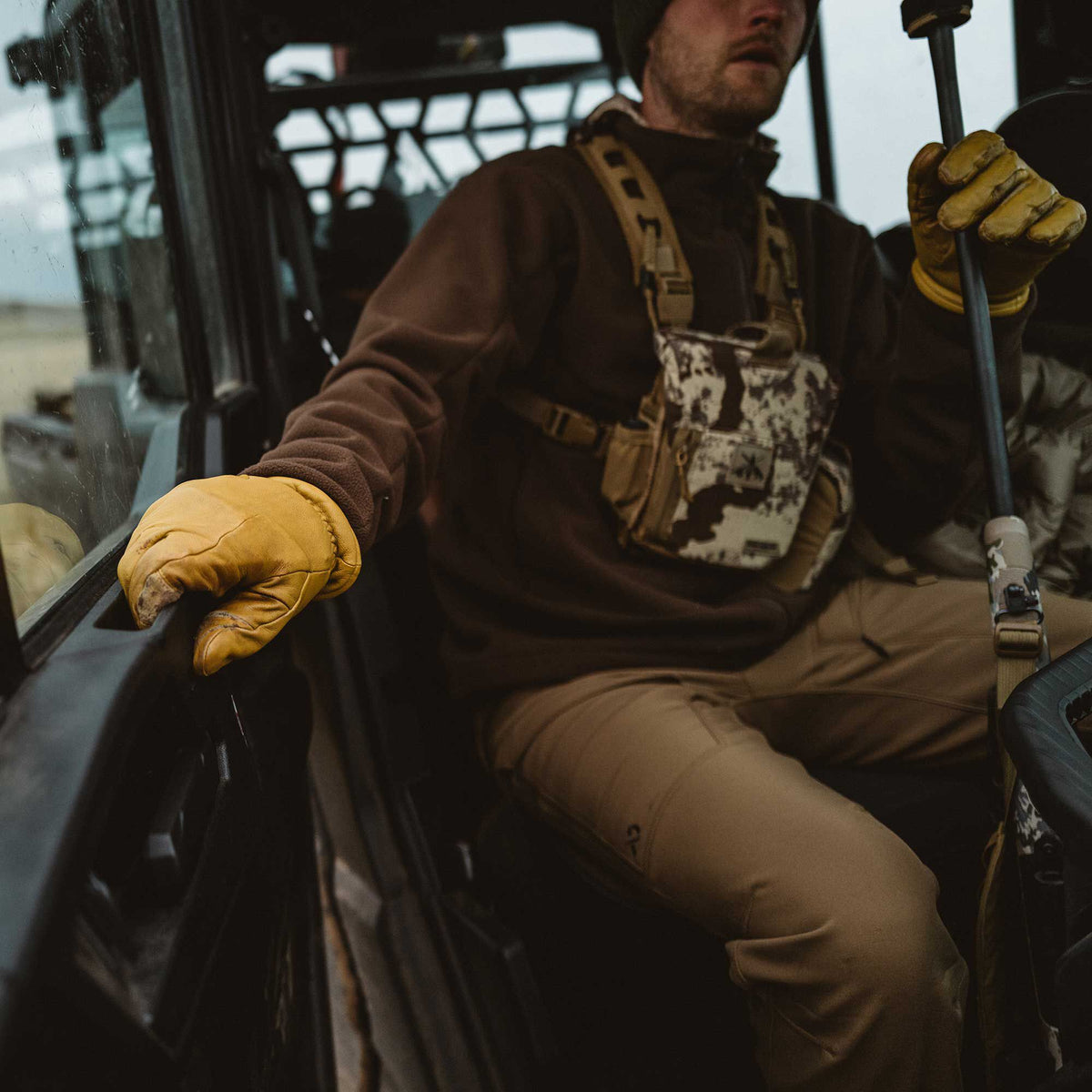 A man wears a brown fleece, tan pants, yellow leather gloves, and a camouflage chest rig while sitting in a vehicle.