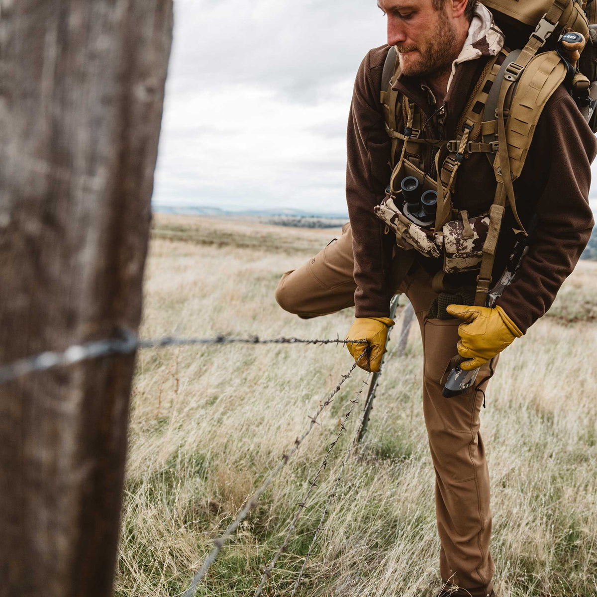 A man wearing a brown fleece jacket, tan outdoor pants, and yellow gloves carefully steps over a barbed wire fence.