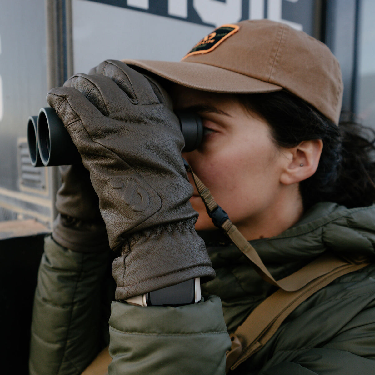 Dark brown textured leather gloves with an embossed logo and elasticized wrist, worn by a person using binoculars, with a smartwatch visible beneath the cuff.