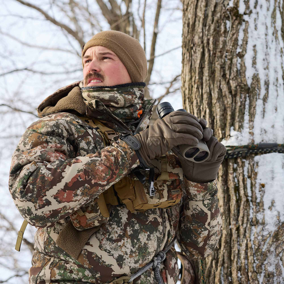 A hunter wearing a brown beanie and multi-toned camouflage jacket with a fleece collar, an FHF GEAR binocular harness, and holding light gray binoculars in a snowy forest.