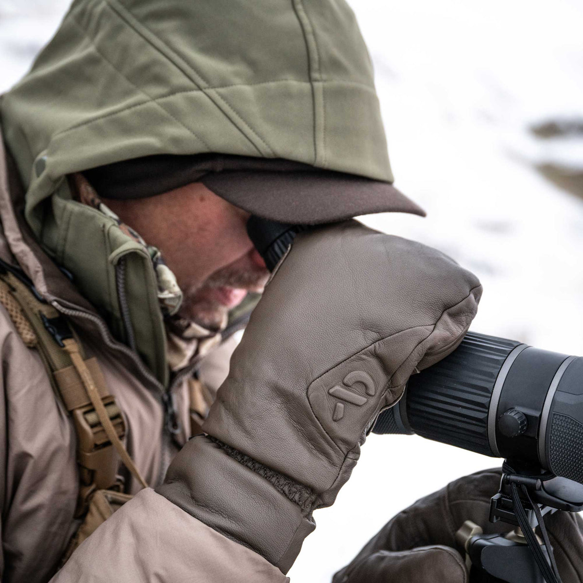 A person wearing dark brown leather mittens with an embossed logo is looking through a spotting scope in a snowy outdoor setting.