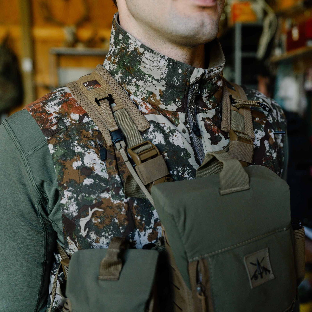 A man wears a camouflage jacket with olive green sleeves and an olive green chest rig with tan harness straps.