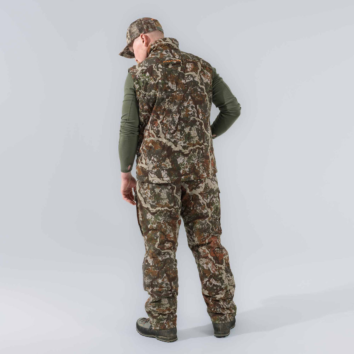 A person from behind wears a First Lite camouflage hunting vest with orange logo, matching pants with leg zippers, an olive green long-sleeve shirt with thumbholes, a camouflage cap, and boots.