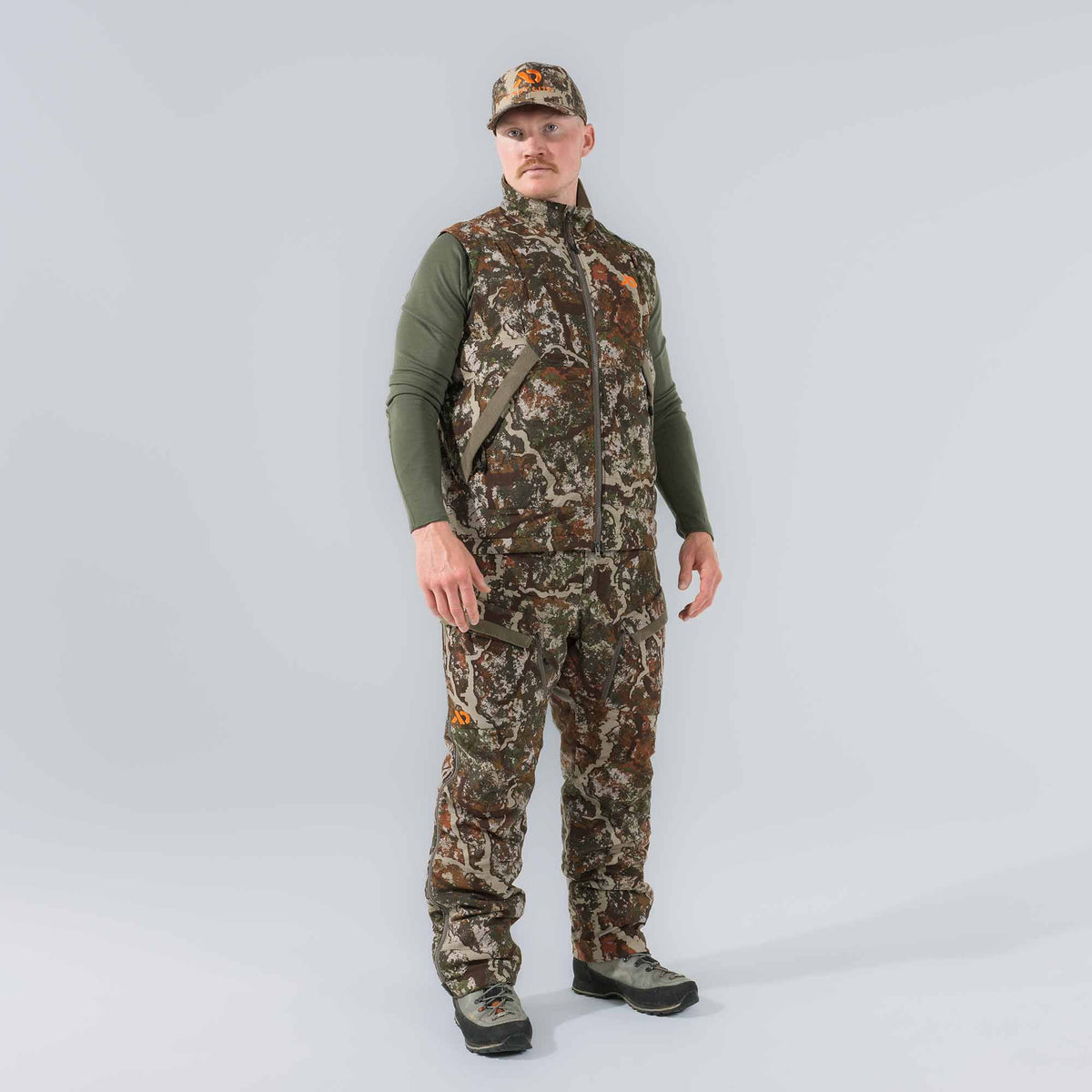 A man in a full camouflage hunting outfit, featuring a vest, pants, and cap in a green, brown, and tan pattern, with orange logos.