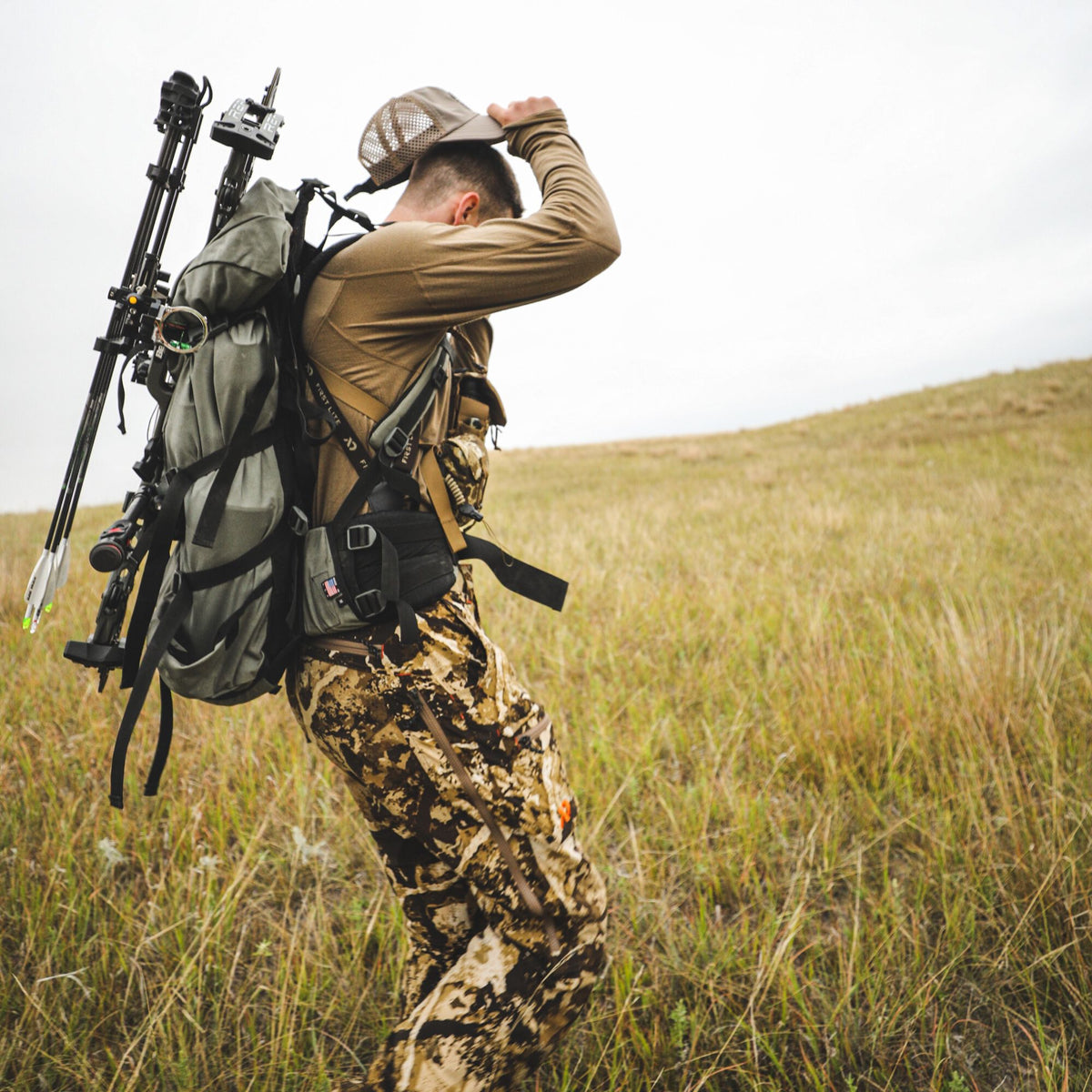 Person wearing a tan long-sleeve shirt, camouflage pants, and a grey hunting backpack with a bow and arrows, walking in a grassy field.