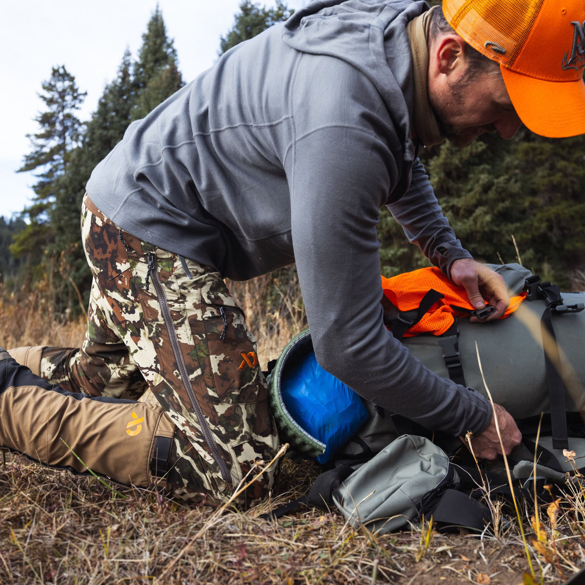 A person wearing a grey seam-detailed hooded shirt and brown camouflage pants with an orange logo and side zippers packs an olive green backpack.
