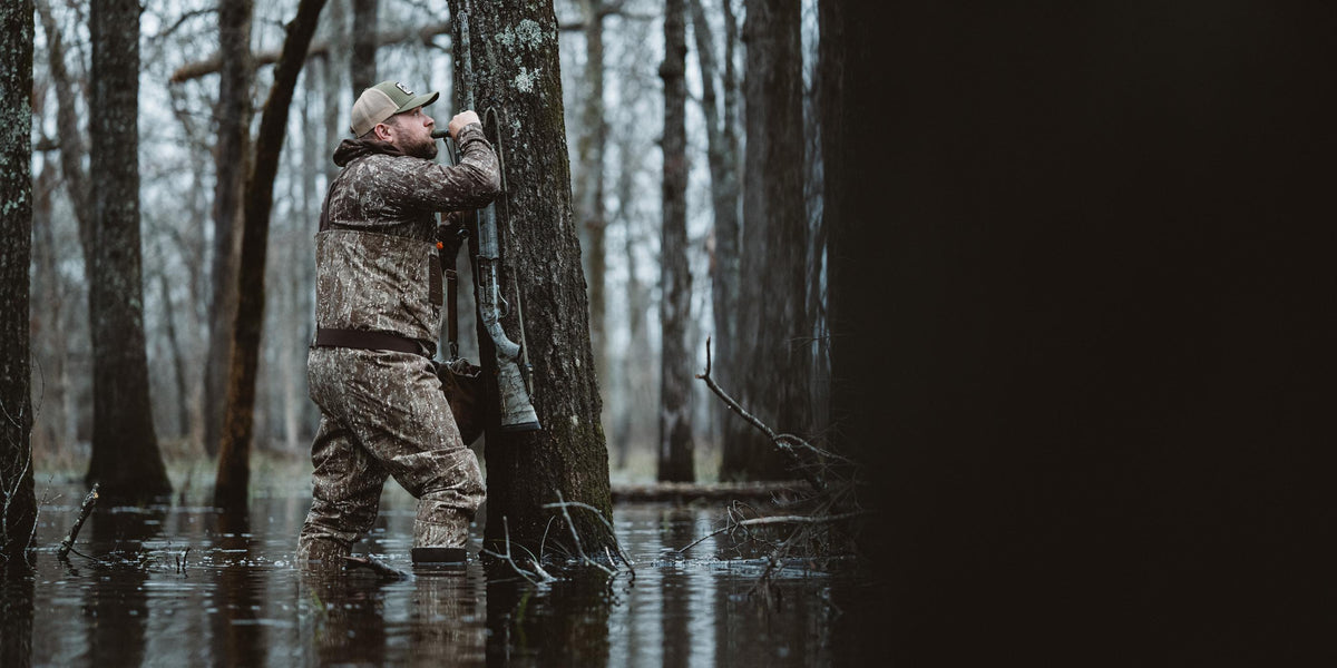 Hunter in camouflage waders uses a duck call in a flooded forest, shotgun beside tree.