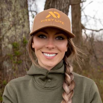 Tan First Lite trucker hat with yellow logo and text on smiling woman.
