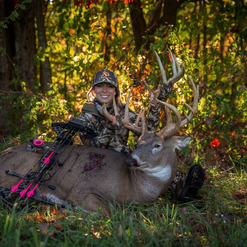 Smiling huntress in camo with harvested buck and a pink-accented compound bow.