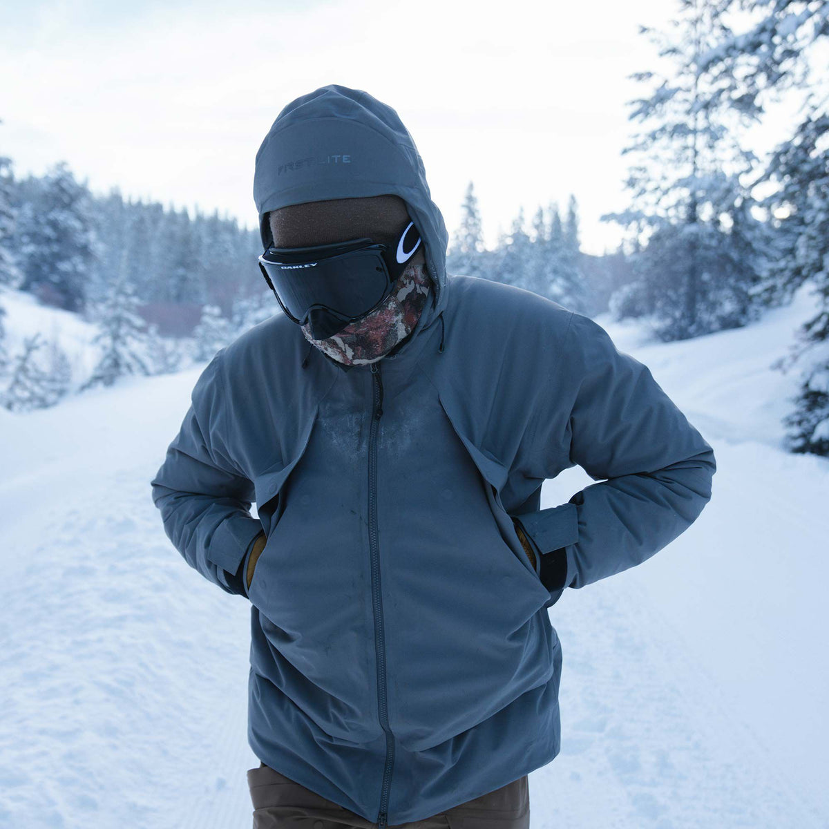 A gray hooded "FIRST LITE" jacket worn by a person with black ski goggles and a camouflage neck gaiter.
