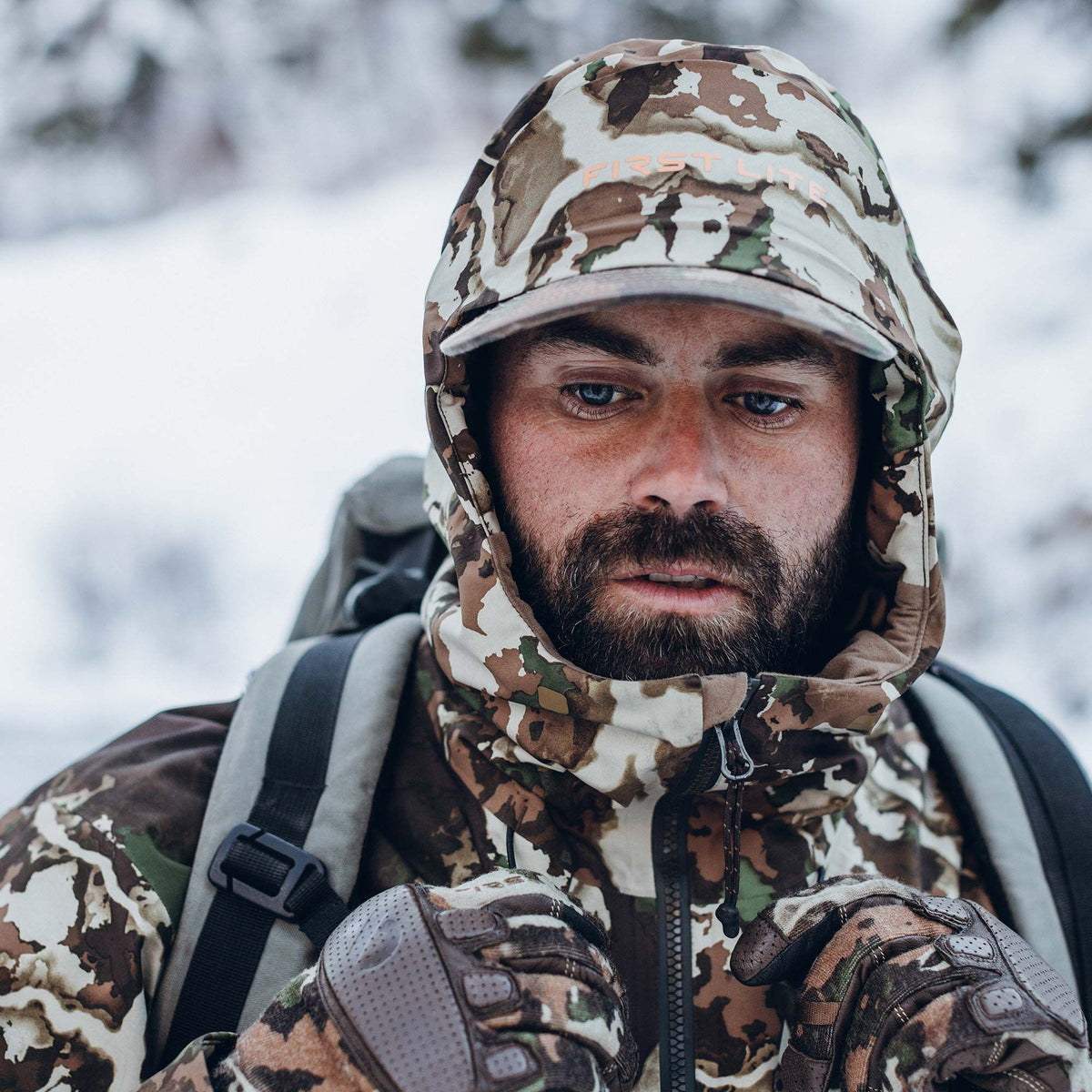 A man wears a First Lite camouflage hooded jacket with a visible logo, matching gloves, and backpack straps in a snowy environment.