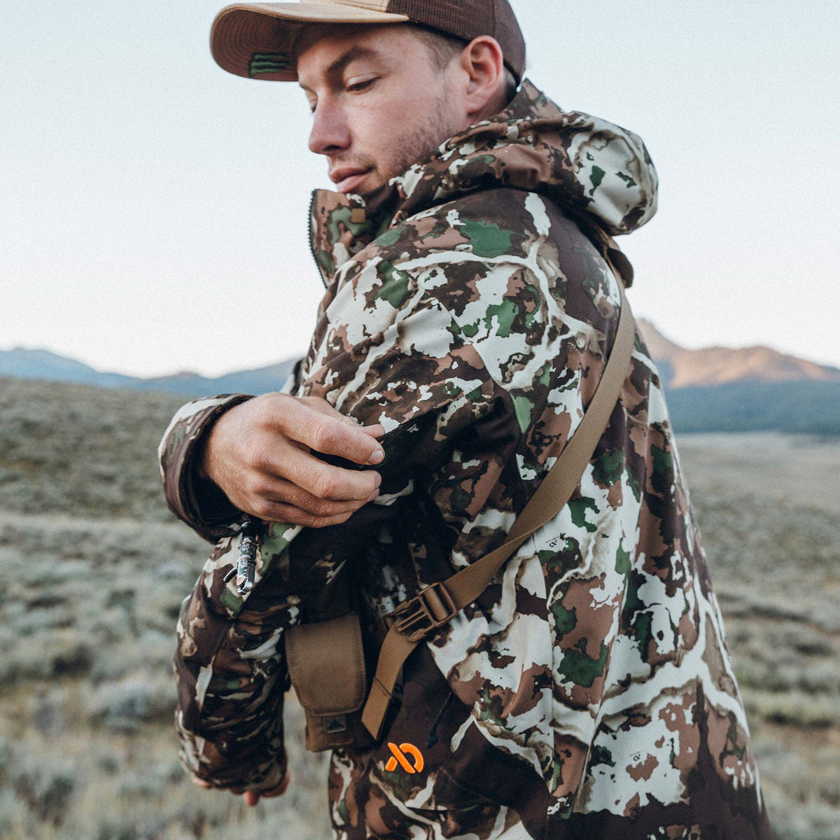 Hooded camouflage jacket in brown, green, and white worn by a man who is adjusting its sleeve, featuring a tan chest strap and orange logo.