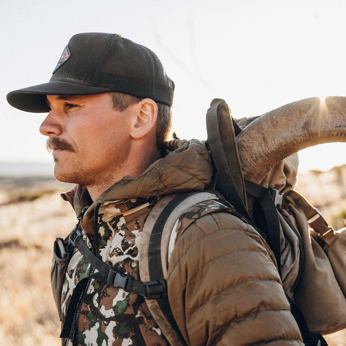 Man wearing a dark grey mesh trucker cap with a patch, a brown quilted jacket, and a backpack carrying a ram horn.
