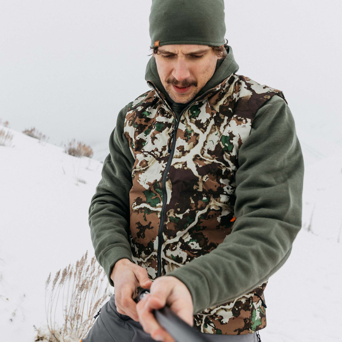 Full-zip camouflage vest worn over an olive green hooded long-sleeve shirt and matching beanie in a snowy outdoor scene.