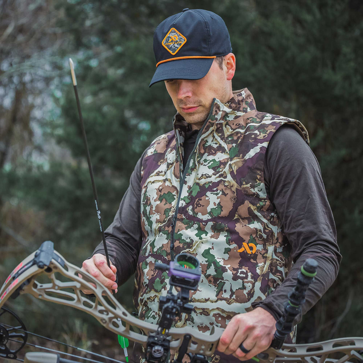 A man in a multi-color camouflage hunting vest and a navy blue cap with an orange brim cord and diamond logo patch holds a bow and arrow.