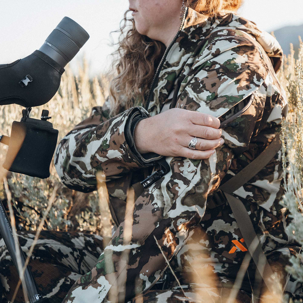 A person wears a brown, green, and white camouflage hunting jacket with zippered pockets and an orange logo while sitting in tall brush next to a spotting scope.