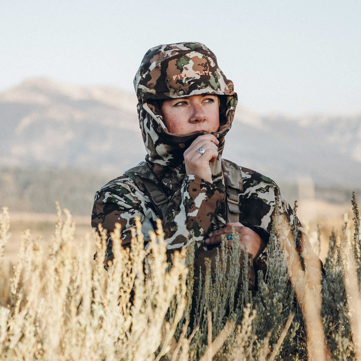 A person wears a brown, green, and tan camouflage hooded hunting top with an orange FIRST LITE logo, adjusting its balaclava-style collar.