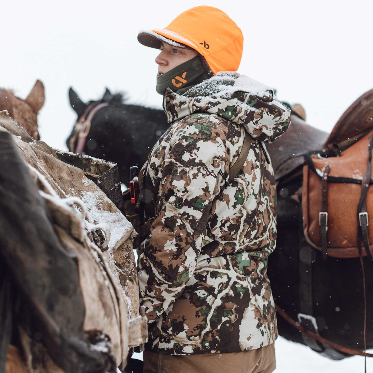 A person wears a white, brown, and green camouflage hunting jacket with an orange cap and green neck gaiter, standing in snow next to a horse.