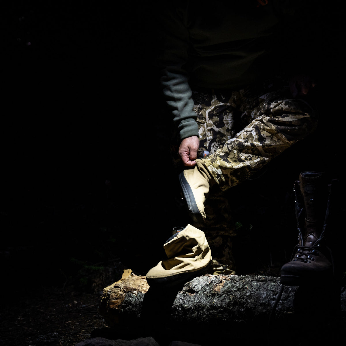 A tan, flexible bootie with a black sole is adjusted by a person, resting on a log next to a dark hiking boot.