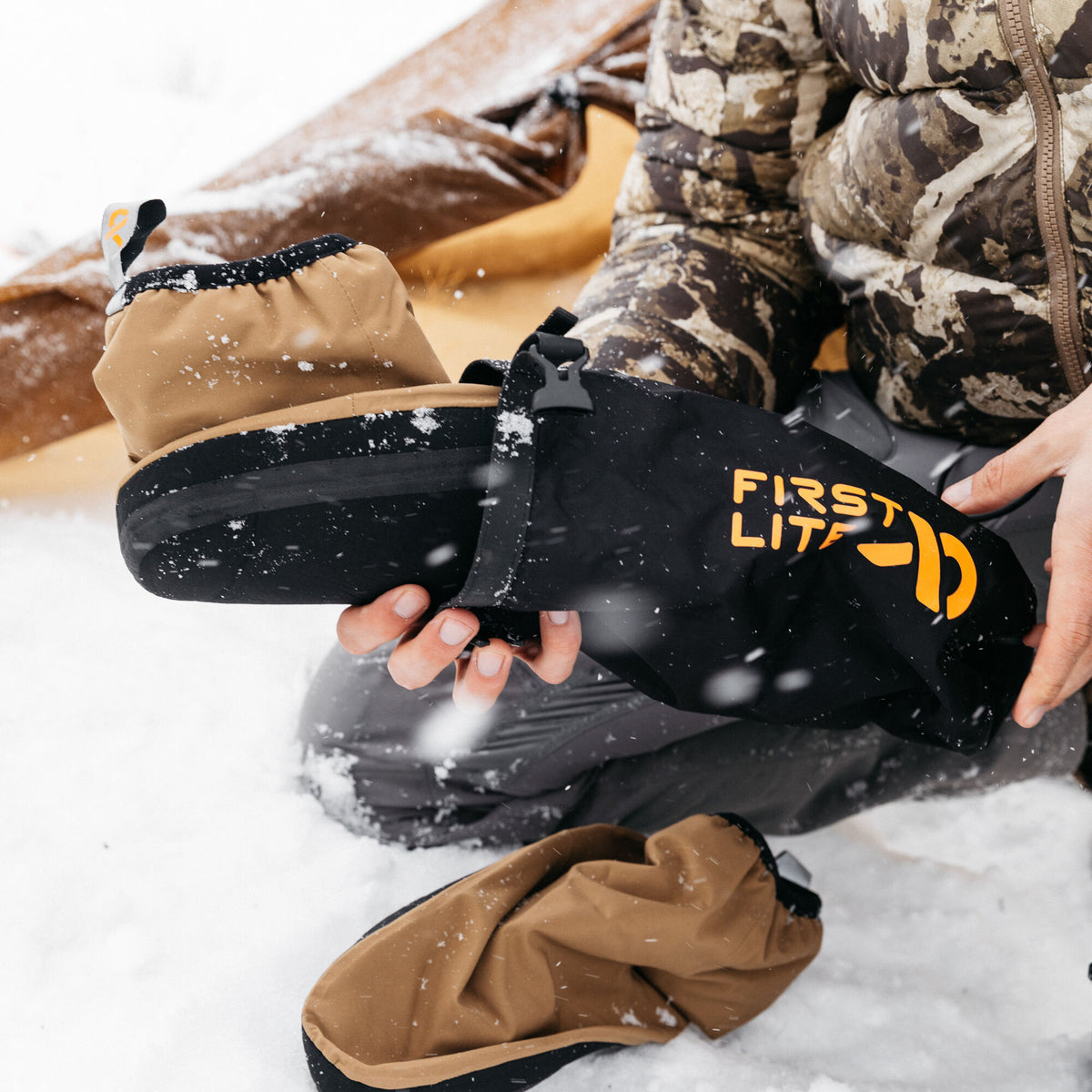 Tan insulated camp bootie with black sole, being placed into a black carrying bag featuring an orange 'FIRST LITE' logo, amidst falling snow.