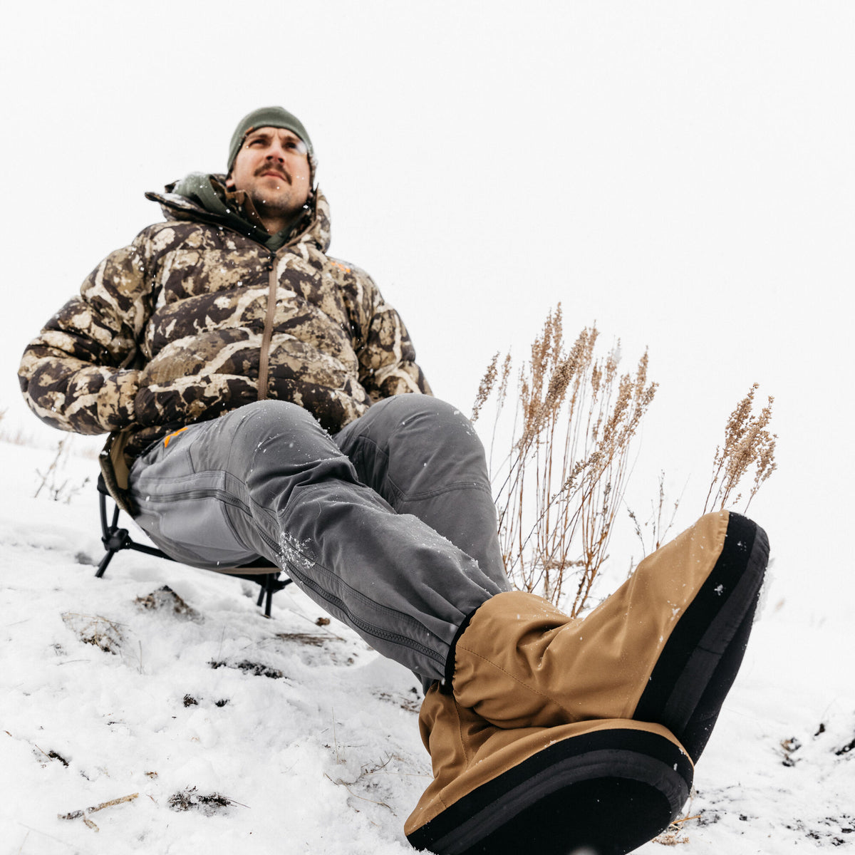 Tan insulated snow boots with thick black soles and a black side stripe worn by a person sitting in the snow.