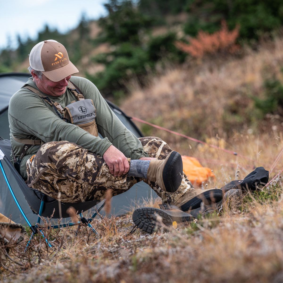 A man wearing a First Lite brown and white trucker hat, olive green shirt, camouflage hunting pants, and a Leupold chest pack adjusts his brown hiking boot over a grey sock in an outdoor camp setting.