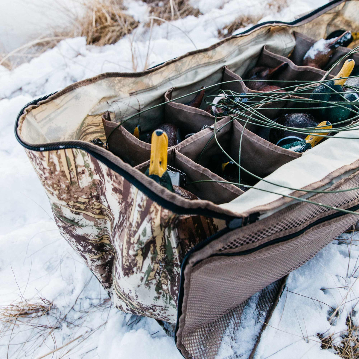 Open camouflage duck decoy bag with internal dividers, containing duck decoys and lines, resting in the snow.